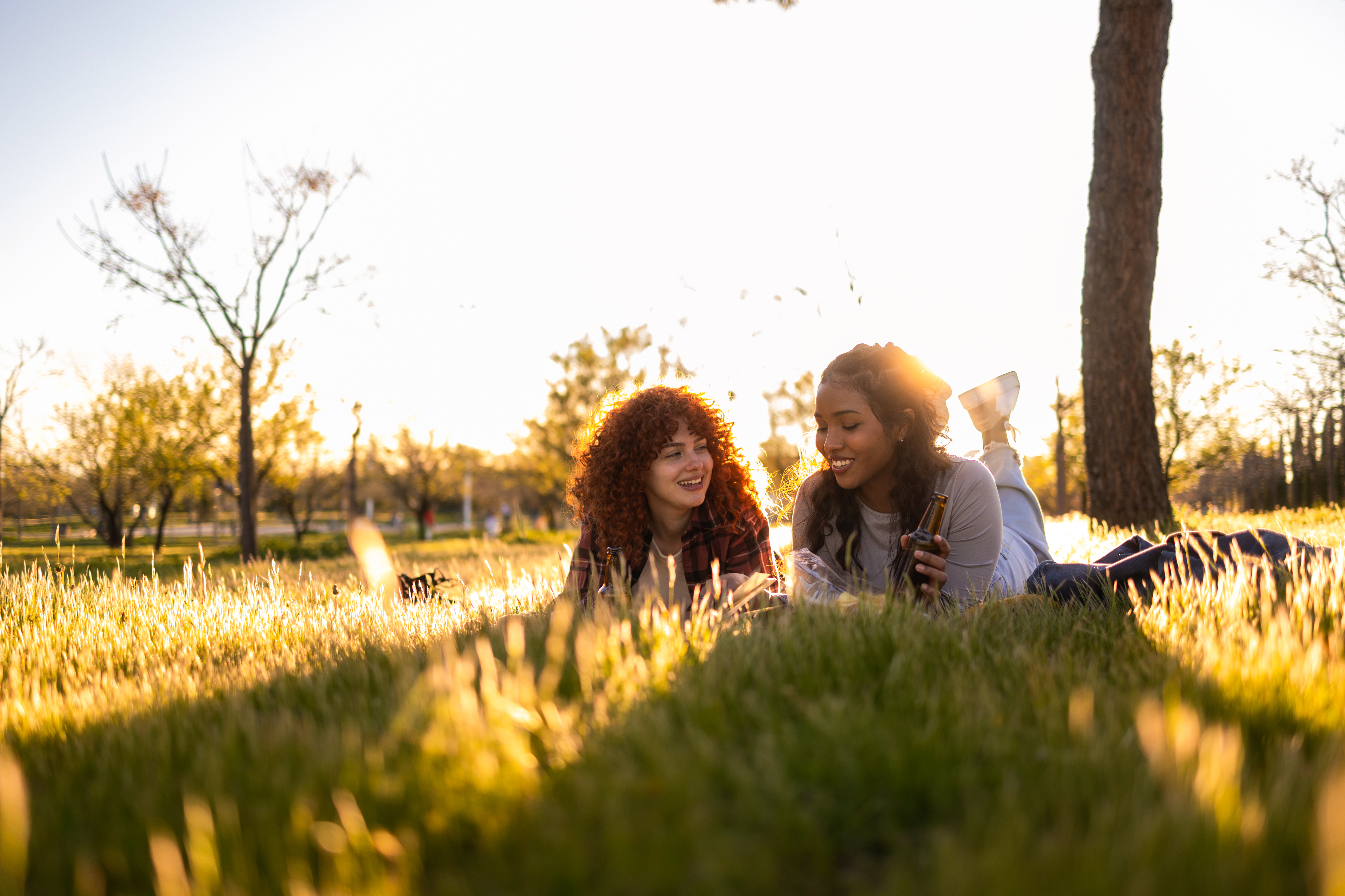 Two women laugh and talk while laying in the grass with the sun setting behind them