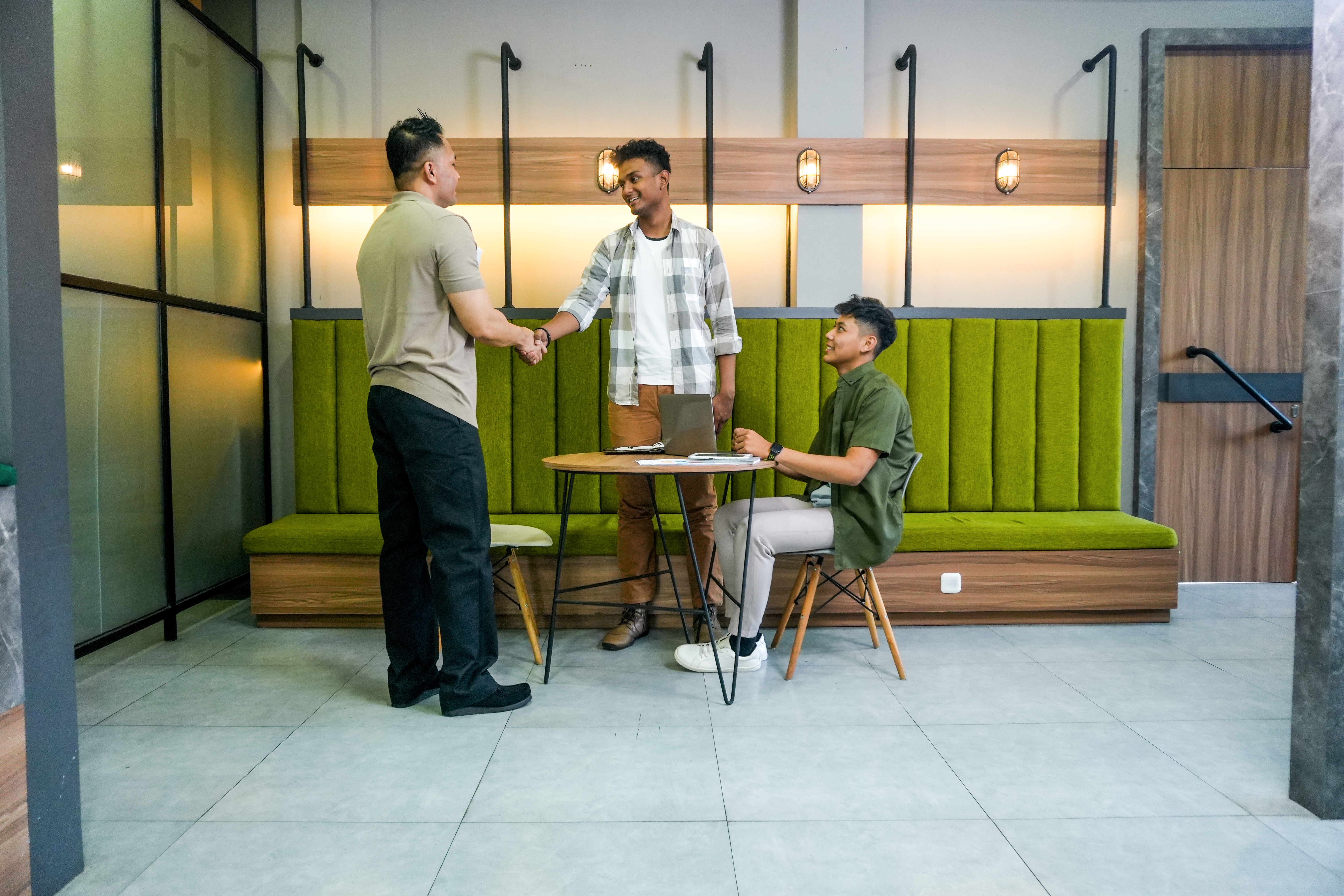 Three people meet over table with papers and a laptop on it, two of them are shaking hands.